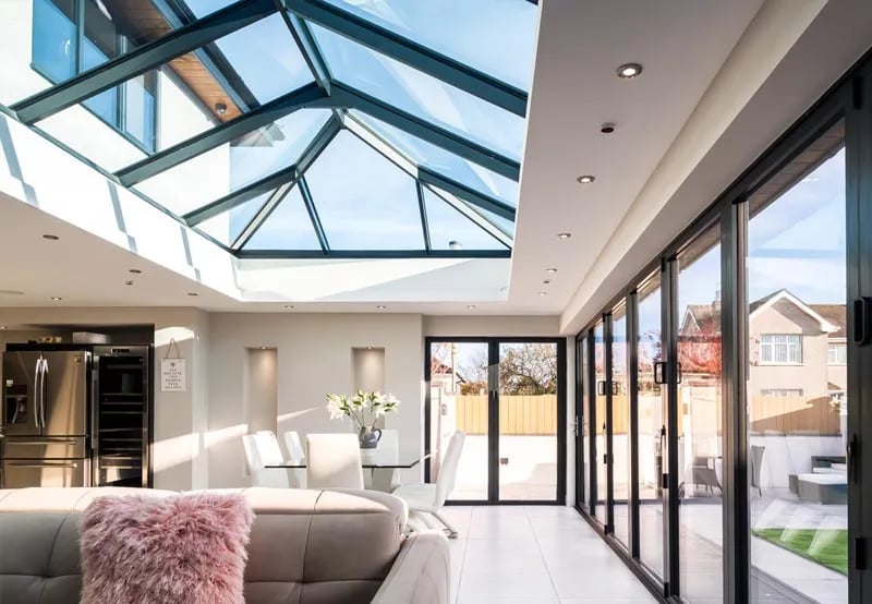 Modern living room with skylight roof, gray sofa, large glass doors opening to patio, and contemporary kitchen