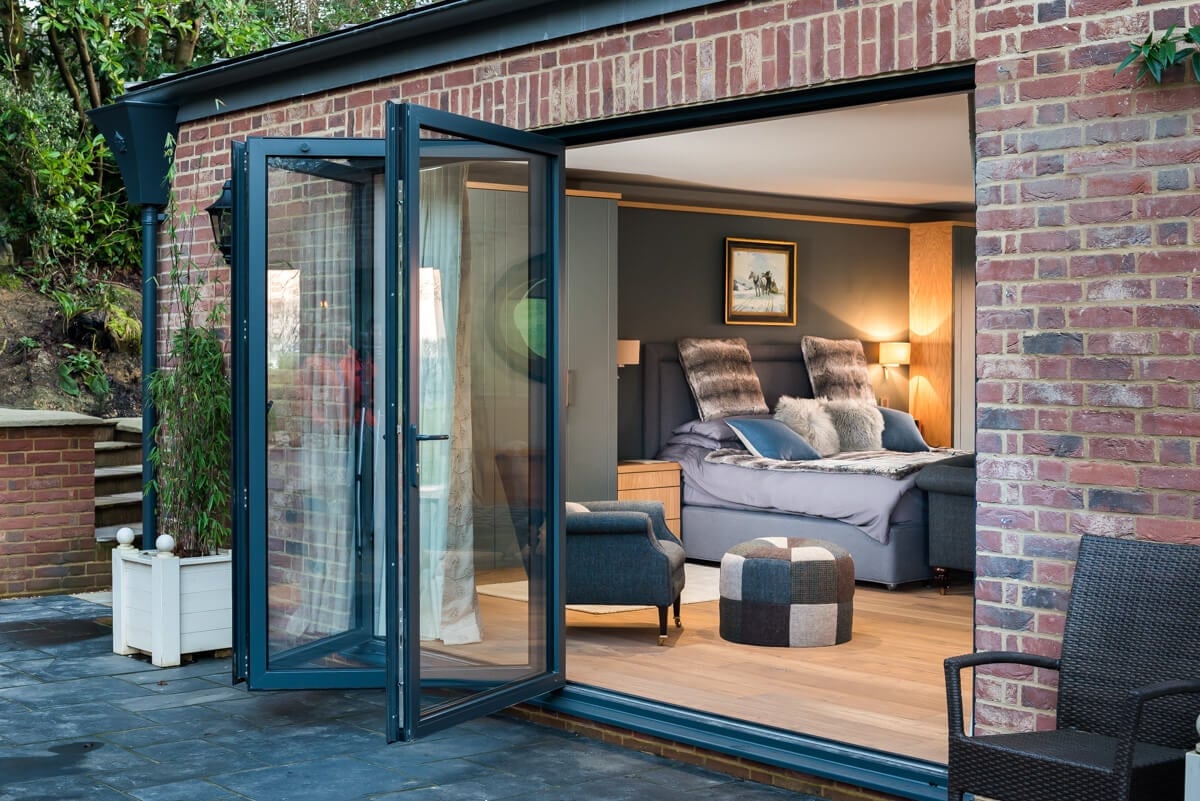 Modern glass bifold doors opening to a contemporary bedroom with bed, warm lighting, and wooden flooring attached to brick exterior
